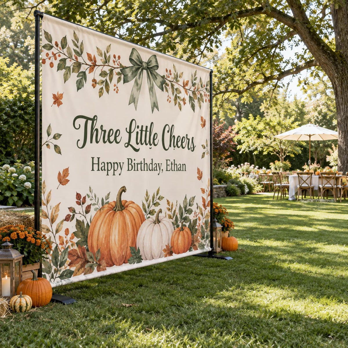 Three Little Cheers birthday backdrop with green bow, pumpkins and autumn leaves in a garden.